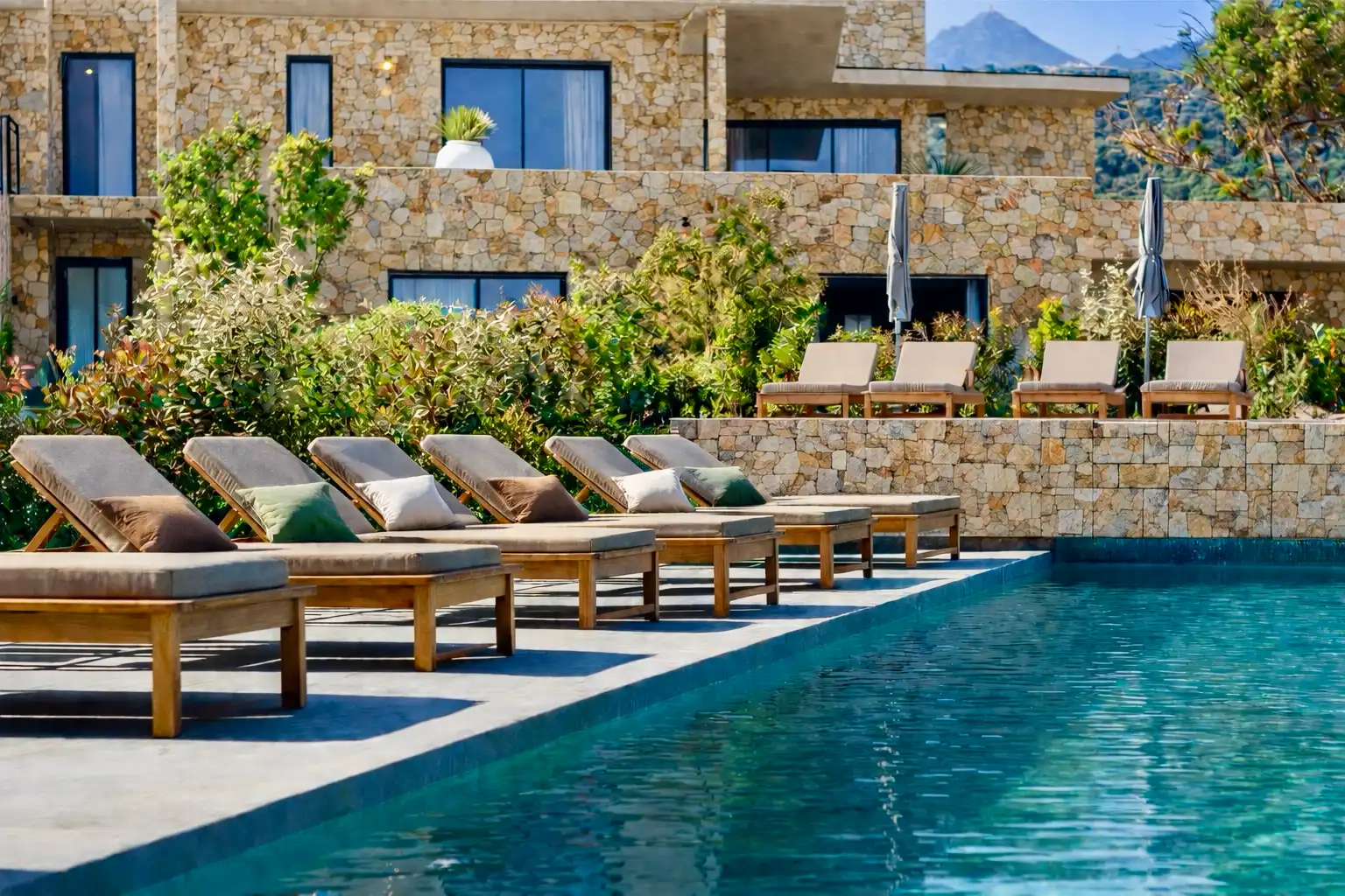 Rangée de bains de soleil en bois avec coussins, alignés au bord d’une piscine ; en arrière-plan, terrasse paysagée et bâtiment en pierre avec parasols.