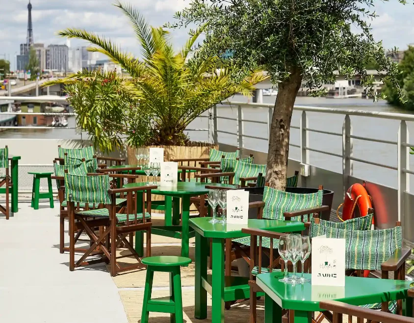 AL24_MAISON_JAUNE_ROOFTOP_DSC6050_WEB Terrasse au bord de l’eau avec tables vertes et chaises à coussins rayés; menus « JAUNE », olivier, palmier et bouée orange; tour Eiffel au loin.