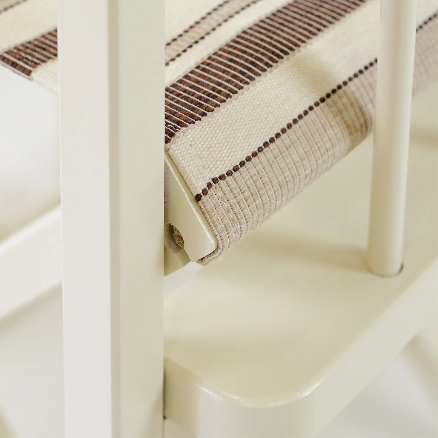 Gros plan d’une assise en toile écrue à rayures brunes, tendue sur un cadre blanc, avec pièce de fixation métallique visible.