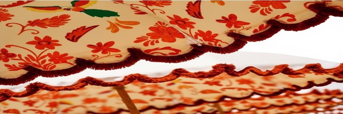 IMG_0303 copie Gros plan sur des parasols en toile beige à motif floral orange et rouge, avec bordures ondulées et franges bordeaux, montés sur des mâts en bois, photographiés en enfilade sur fond clair.