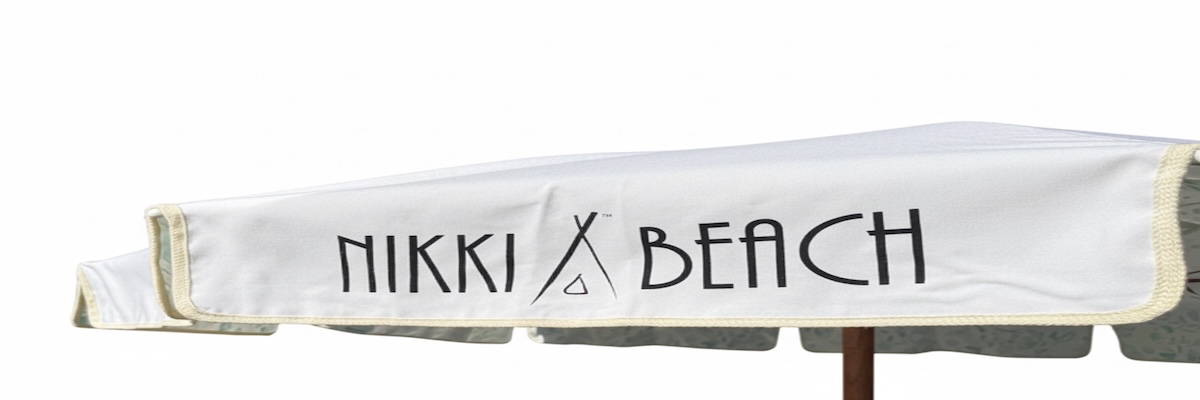 Logotisation Parasol rectangulaire en toile blanche avec l’inscription « NIKKI BEACH » en lettres noires sur le pan latéral, bordures claires et mât en bois, photographié sur fond neutre.