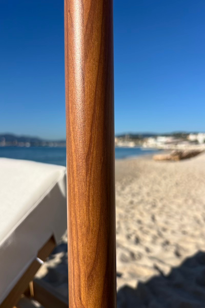 Mât effet bois Gros plan sur un mât de parasol en bois verni, avec en arrière-plan flou une plage de sable, la mer et un ciel bleu dégagé.