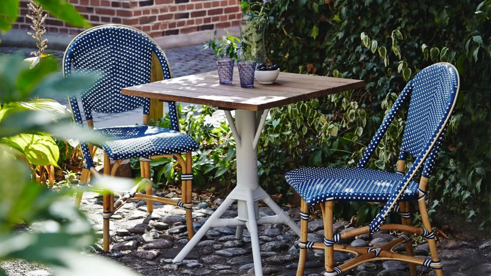 Petite table de bistrot en extérieur avec deux chaises en rotin bleu, sur un sol pavé, entourée de plantes.