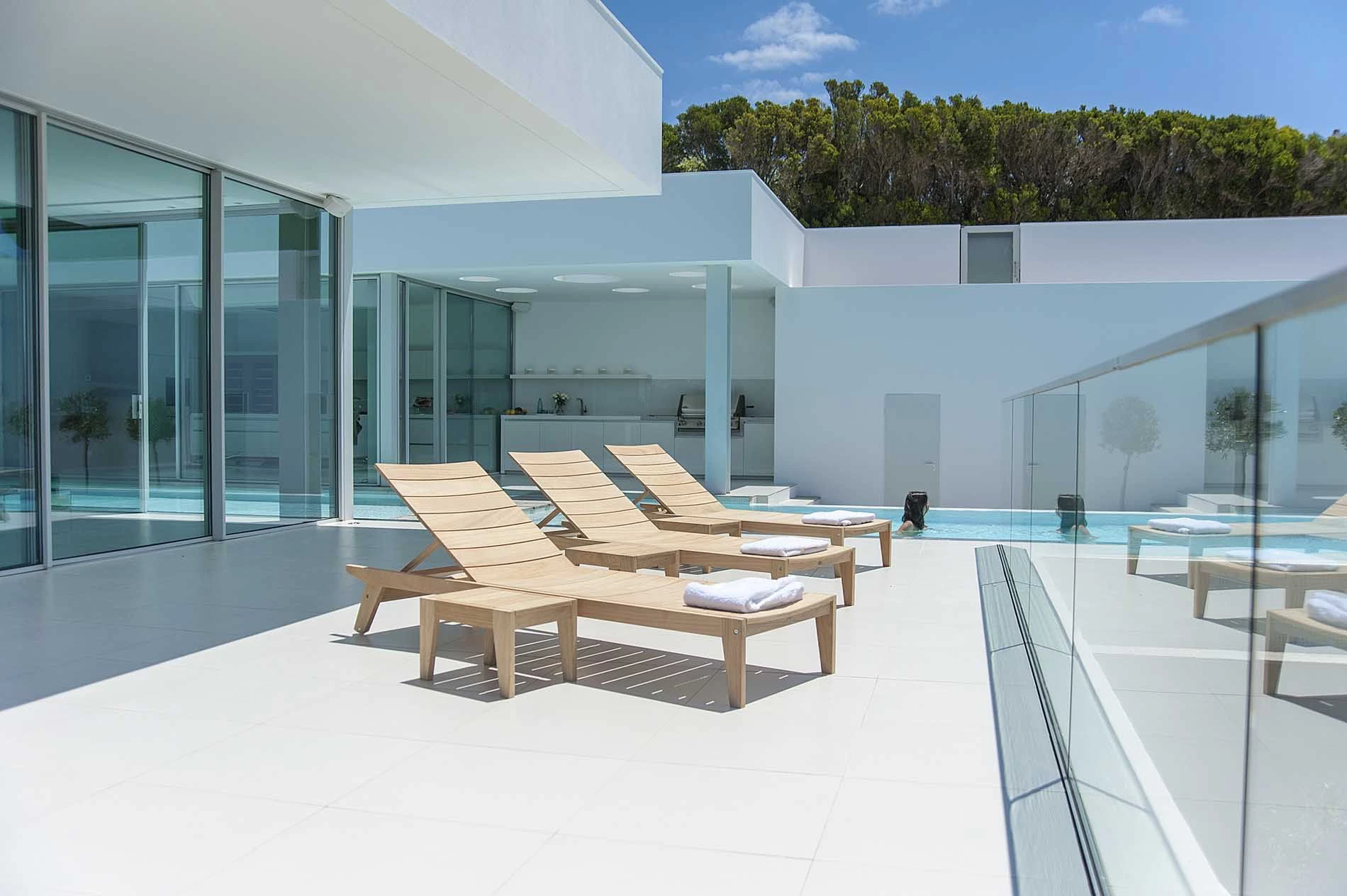 Bains de soleil en bois clair au bord d’une piscine sur terrasse contemporaine avec garde-corps vitré.