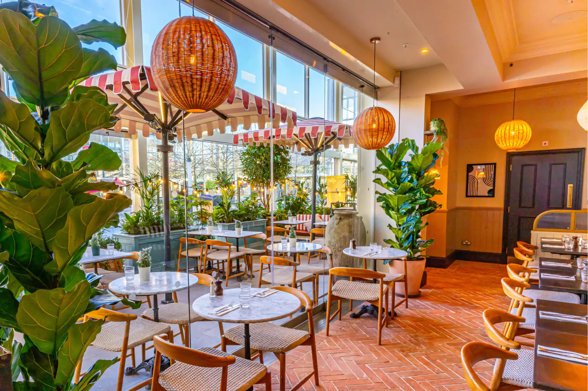 Terrasse de restaurant aménagée avec fauteuils en bois, assises tressées, tables rondes et plantes vertes.