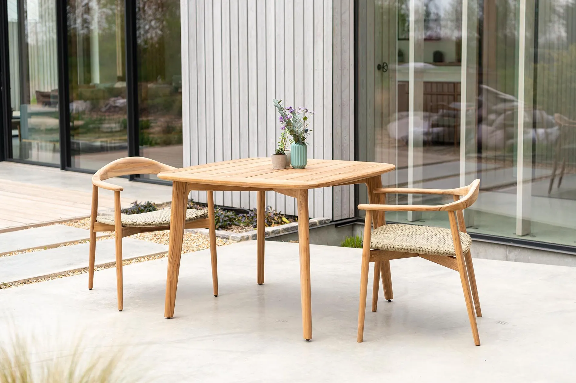 Table carrée et fauteuils de jardin en bois avec assises tressées sur terrasse moderne vitrée.