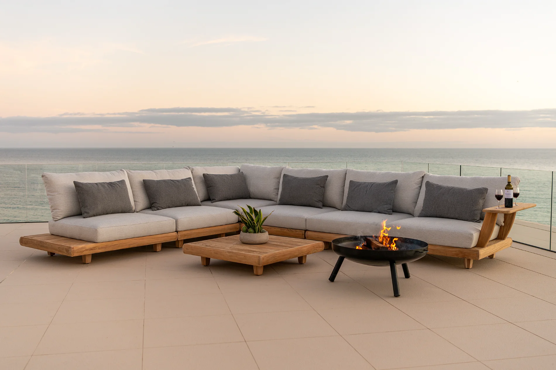 Salon de jardin en bois avec canapé d’angle, coussins clairs et brasero sur terrasse face à la mer.